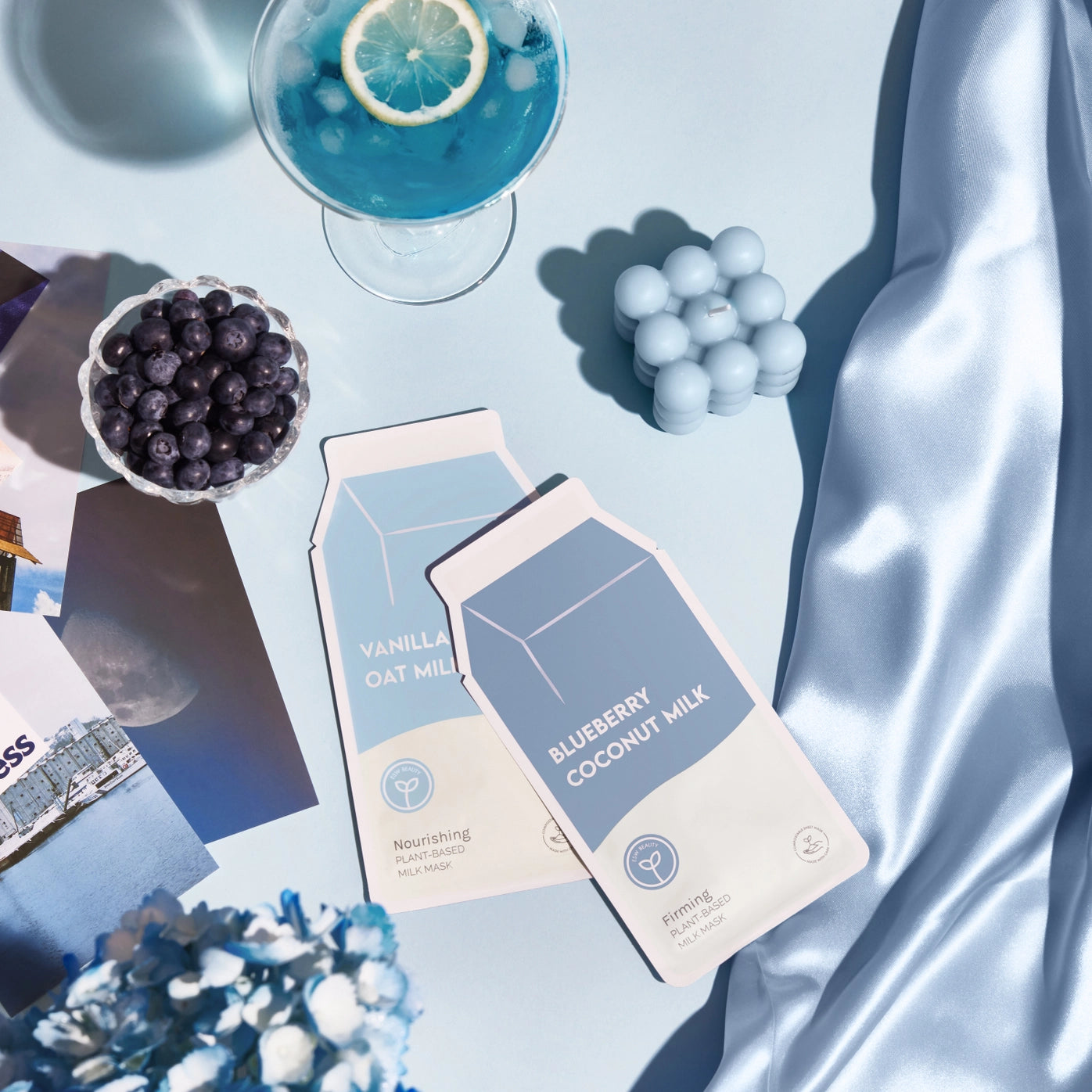 Two cartons of blueberry coconut milk mask on a light blue fabric background with a glass of blue drink and fruit.